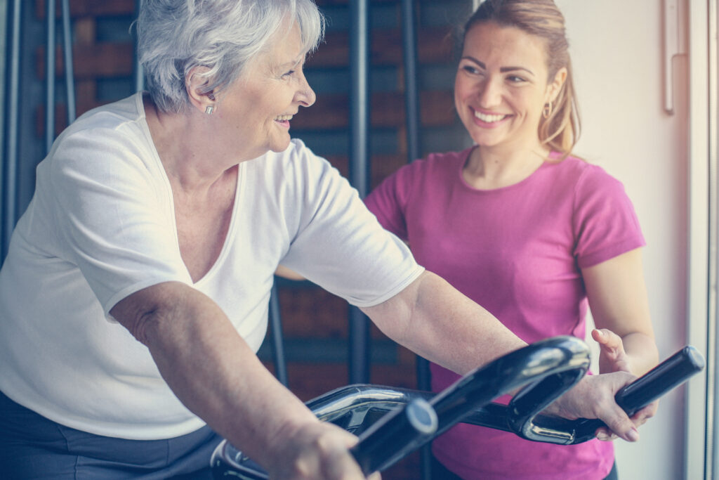 A senior woman pedaling on a bike with a supportive physical therapist beside her, encouraging her during a physical therapy session focused on fitness and mobility