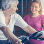 A senior woman pedaling on a bike with a supportive physical therapist beside her, encouraging her during a physical therapy session focused on fitness and mobility
